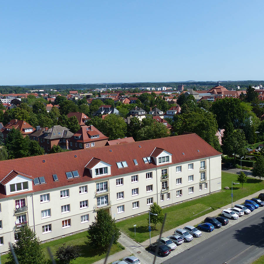 Das Foto zeigt den Blick auf die Carl-Moltmann-Straße. Zu sehen ist ein Mehrfamilienhaus mit rotem Satteldach in grüner Wohnsiedlung, umgeben von Bäumen und weiteren Wohnhäusern, mit geparkten Autos entlang der Straße.