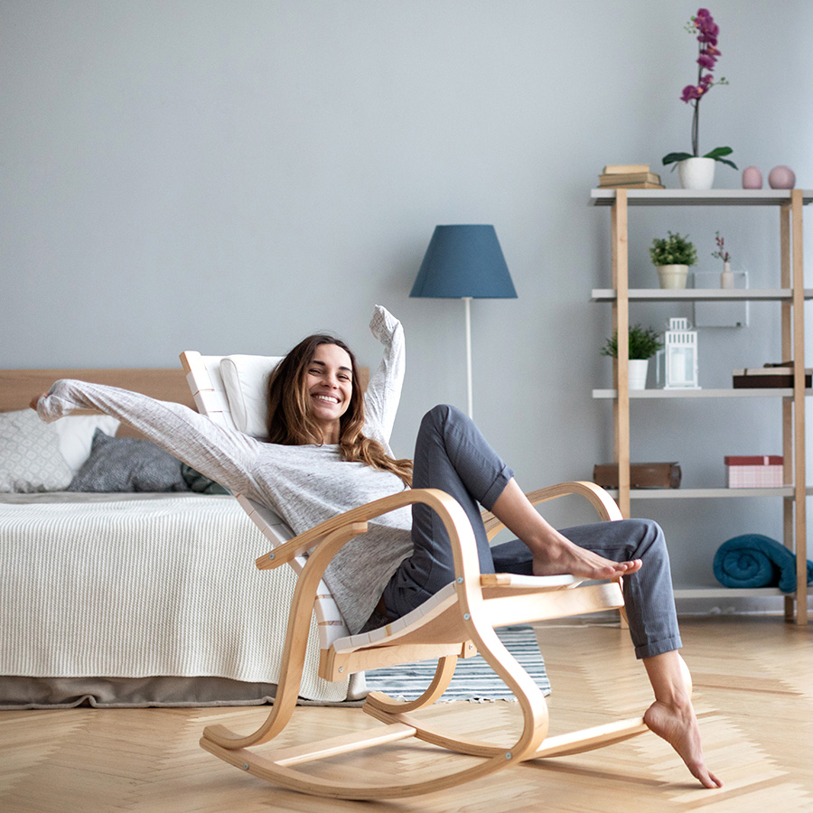 Eine junge Frau sitzt lächelnd in einem modernen Schaukelstuhl in einem gemütlichen, stilvoll eingerichteten Zimmer. Sie trägt bequeme Freizeitkleidung und genießt den Moment der Ruhe.