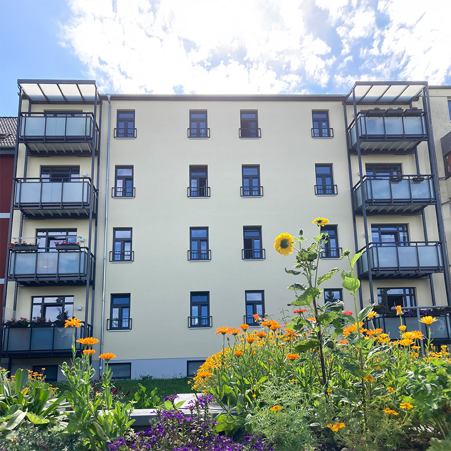 Modernisiertes Mehrfamilienhaus mit Balkonen und heller Fassade, davor blühender Garten mit Sonnenblumen und anderen Sommerblumen unter blauem Himmel.