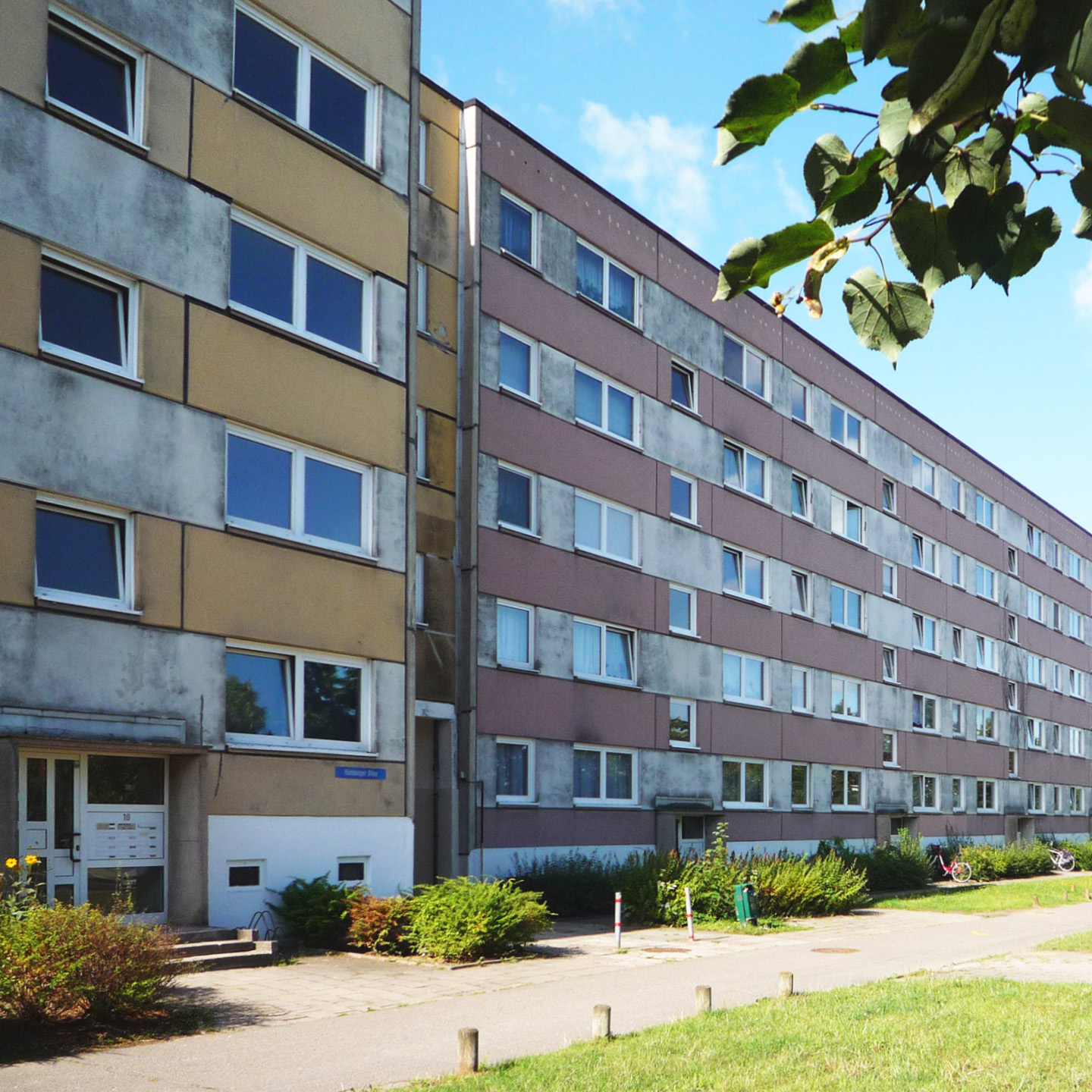 Mehrgeschossiger Wohnblock mit Putzfassade und Balkonen, umgeben von Grünflächen und Gehwegen in einer Wohnsiedlung.