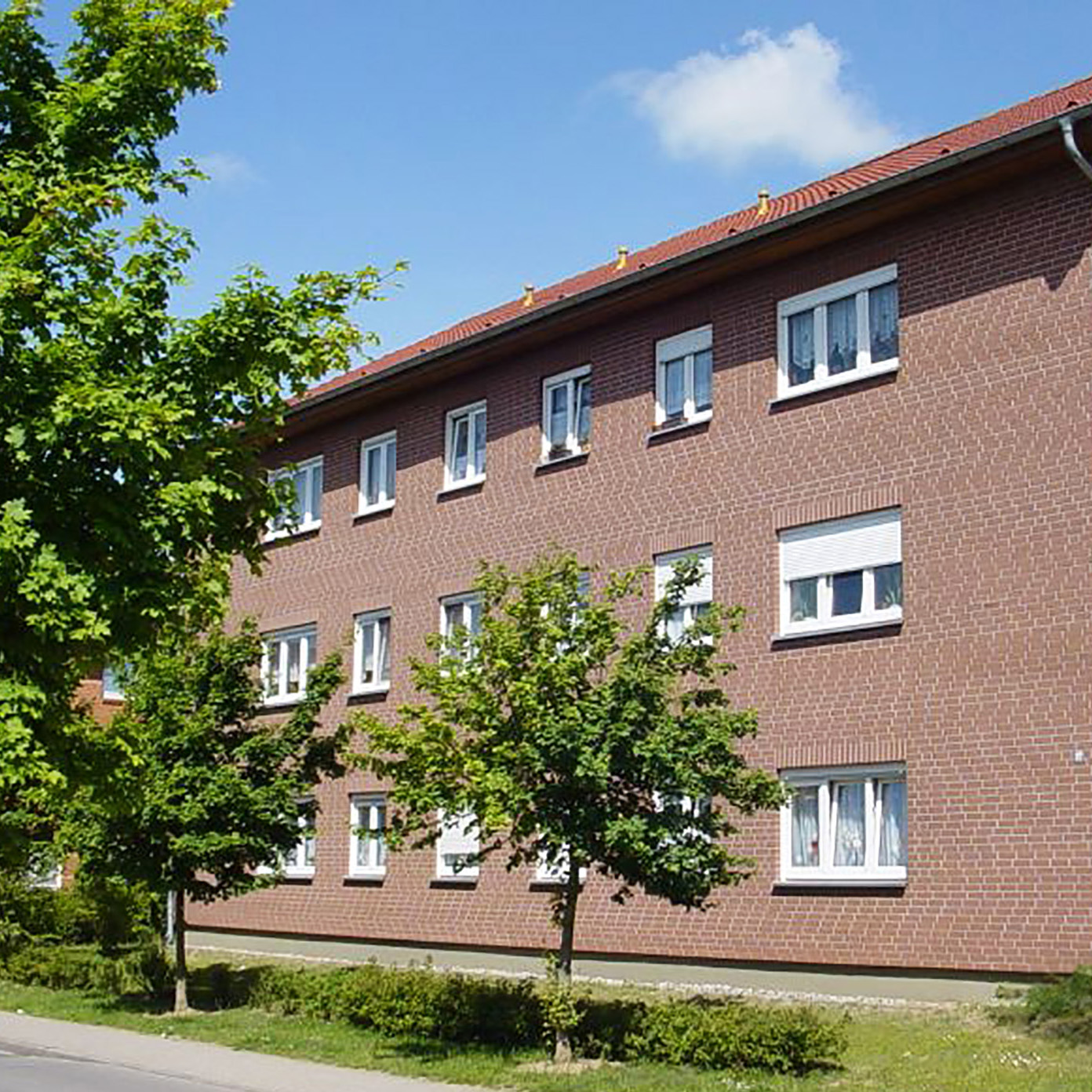 Dreigeschossiges Mehrfamilienhaus mit roter Klinkerfassade und Satteldach, umgeben von Grünflächen und Bäumen, an einer ruhigen Wohnstraße gelegen.