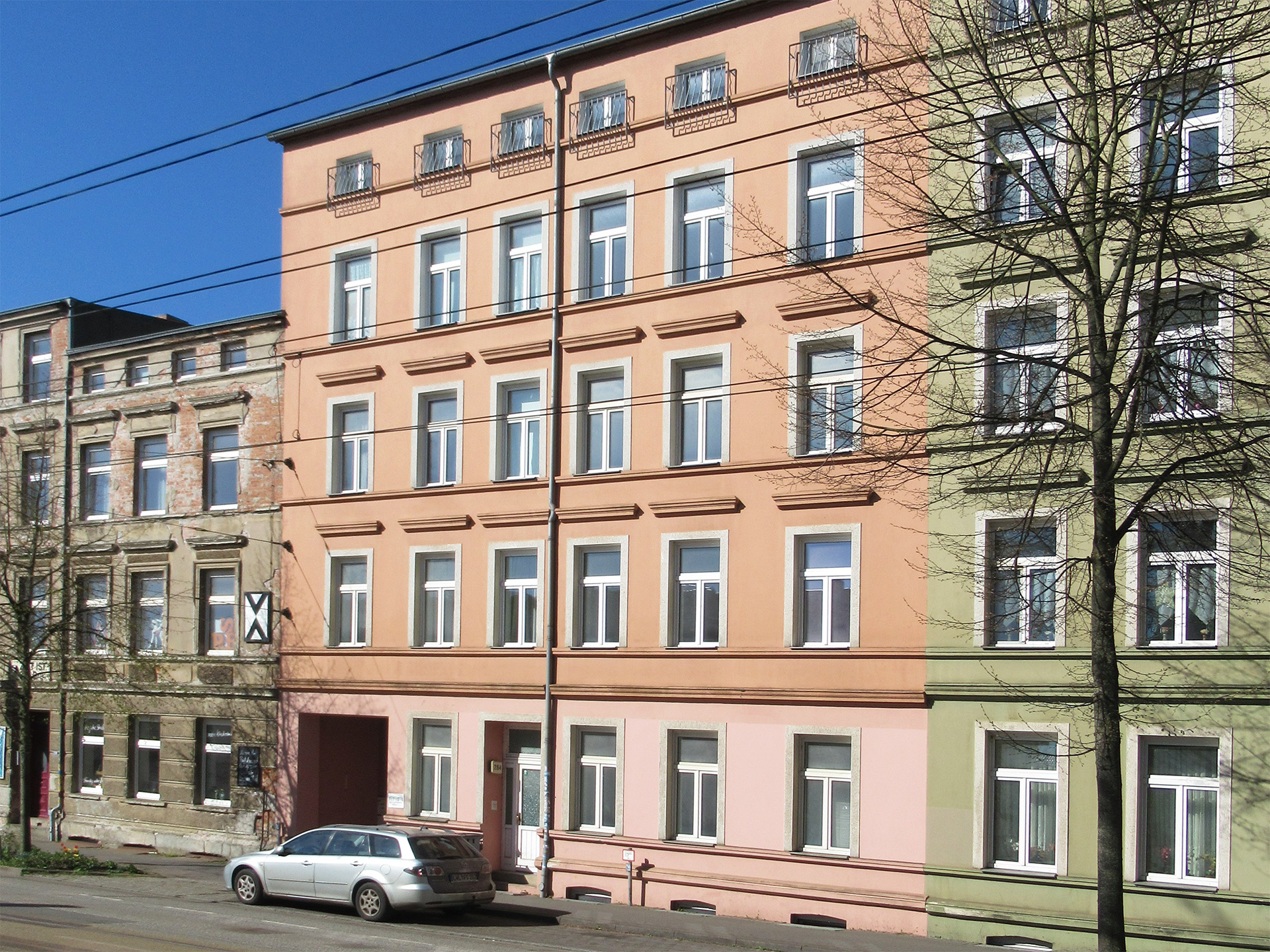 Das Bild zeigt das Wohngebäude in der Wismarschen Straße 284. Es handelt sich um einen Altbau mit hellorangefarbener Fassade zwischen einem unsanierten Nachbargebäude und einem grün gestrichenen Wohnhaus.
