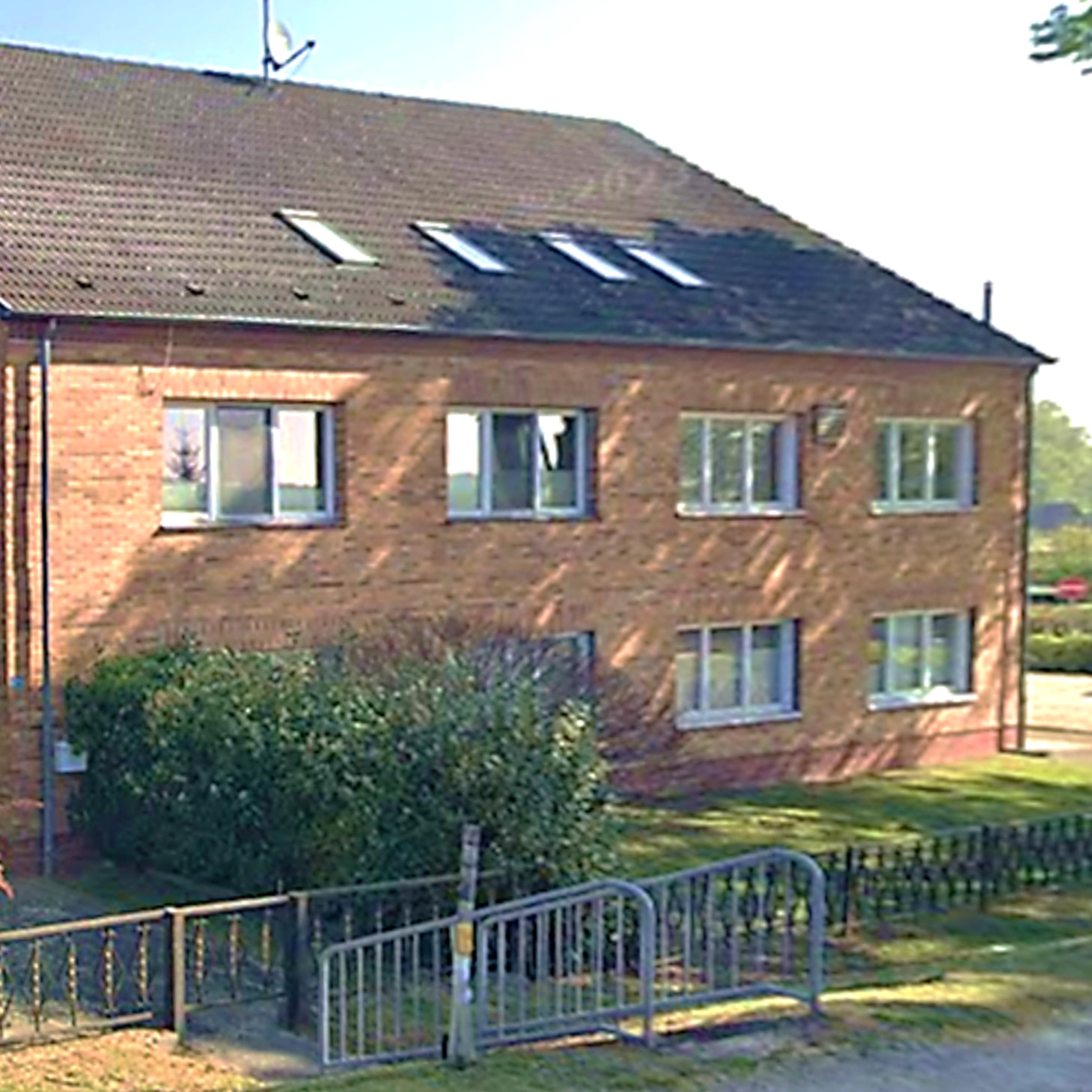 Backstein-Mehrfamilienhaus mit ausgebautem Dachgeschoss, umgeben von Grünflächen und niedriger Einzäunung.