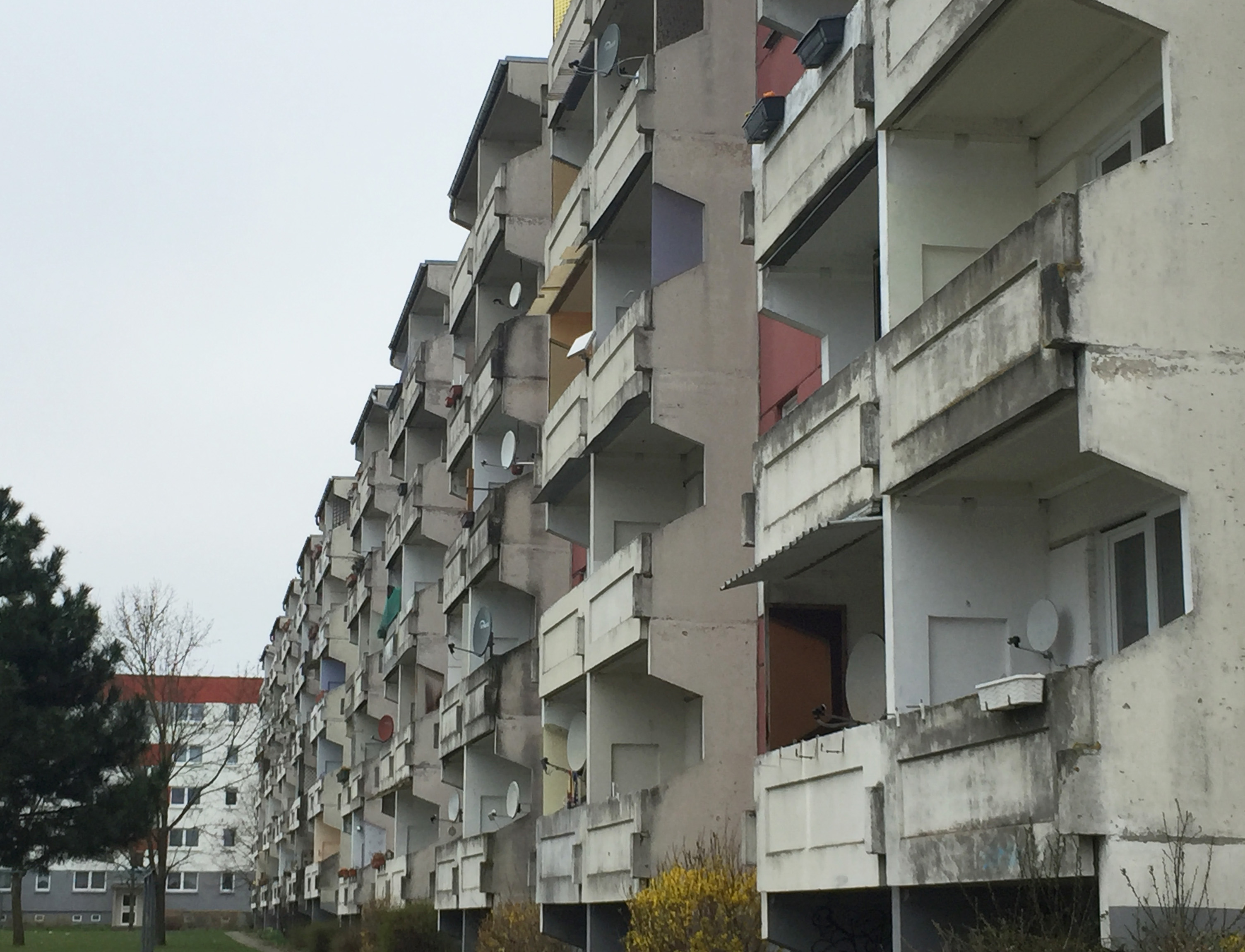 Mehrgeschossiger Plattenbau mit stark verwitterten Balkonen und sanierungsbedürftiger Sichtbetonfassade, aufgenommen aus seitlicher Perspektive.