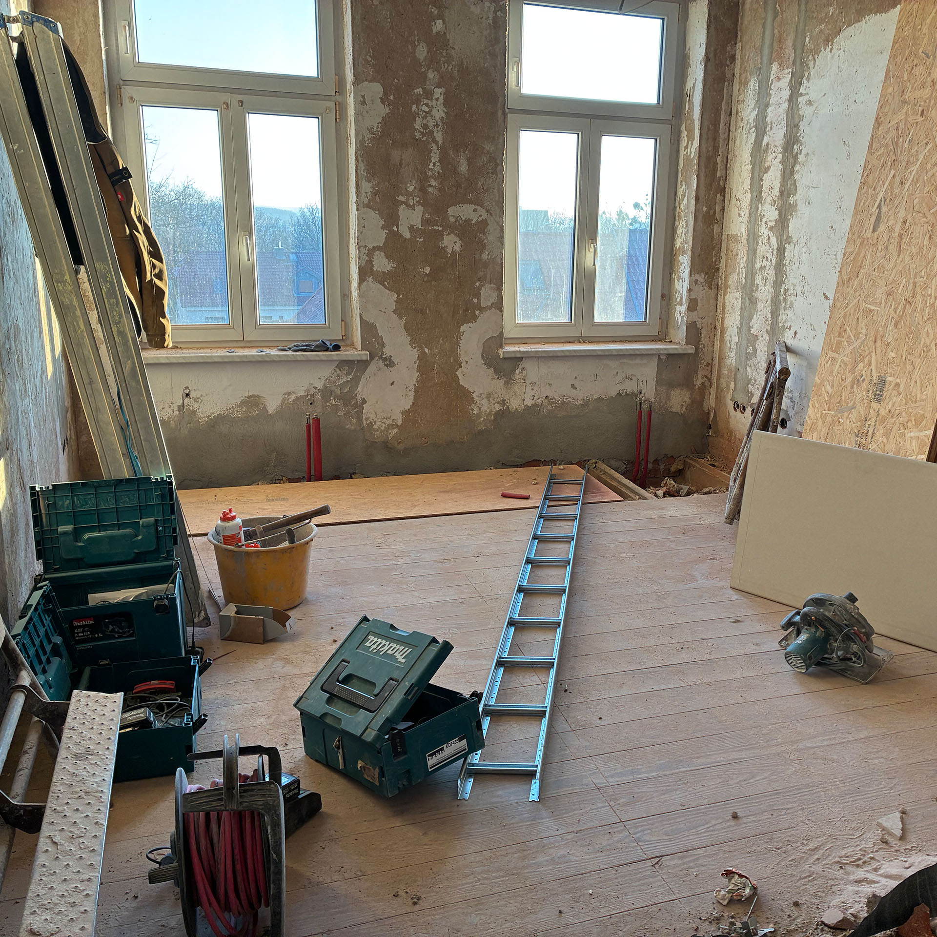 Innenraum einer Wohnung während der Sanierung mit freigelegten Wänden, zwei Fenstern und Bauwerkzeugen wie Leiter, Werkzeugkisten und Kabeltrommel auf dem Boden.