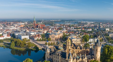 Luftaufnahme der Stadt Schwerin mit dem Schweriner Schloss im Vordergrund, umgeben von Seen, historischen Gebäuden und dem Stadtkern bei klarem Wetter.
